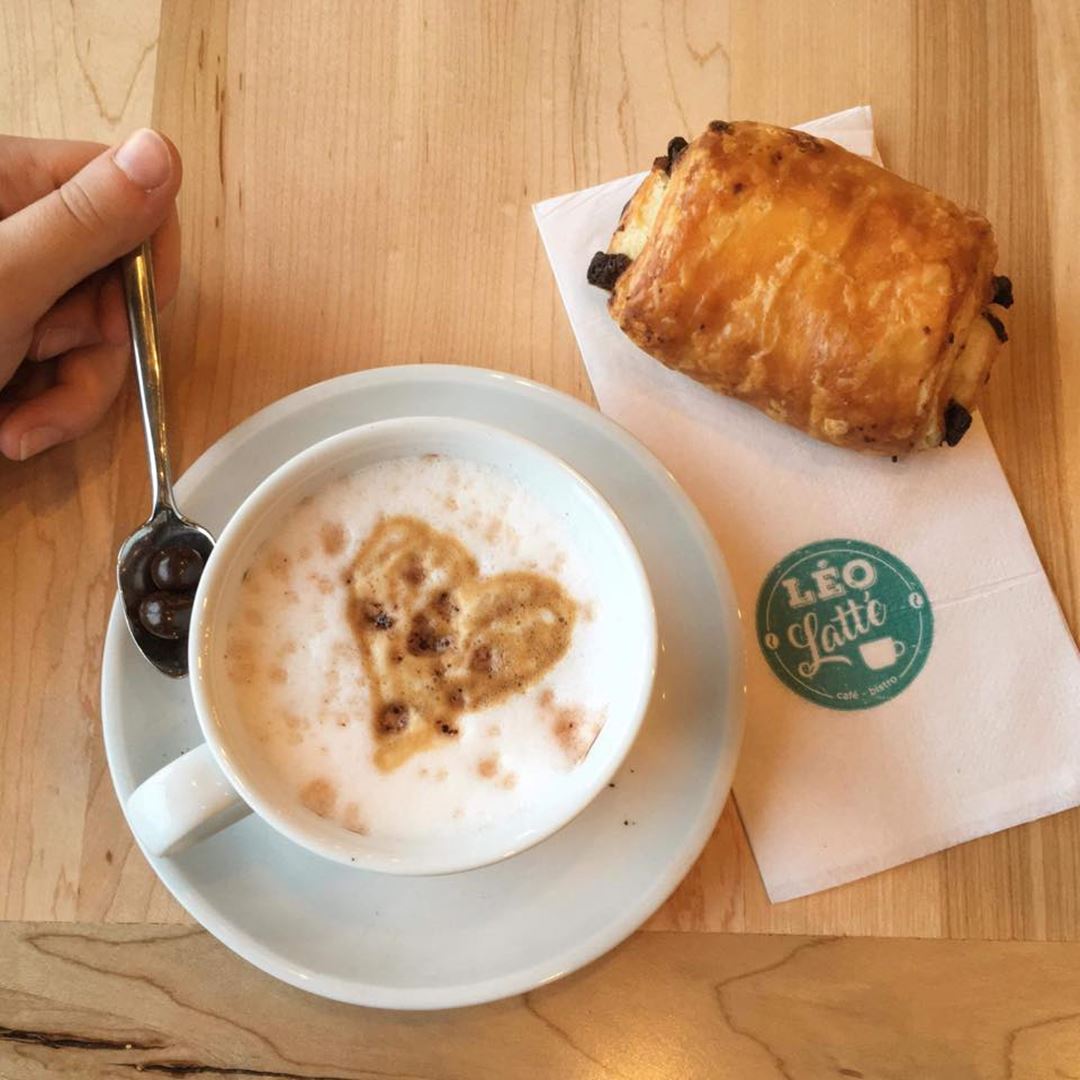 Coffee served in a cup with saucer on which rest a spoon held by a hand accompanied by a chocolatine presented on a counter of blond wood.